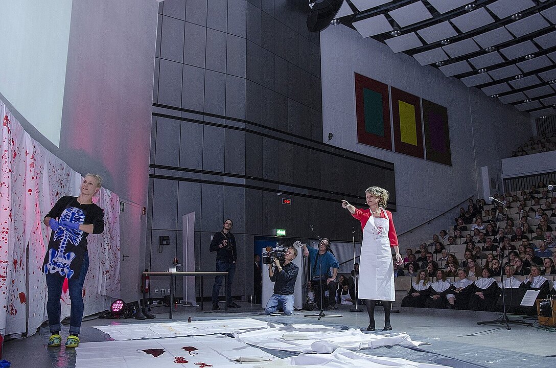 Zwei Personen in einem Hörsaal vorn, eine zeigt auf eine Wand, an der rot bespritzte Tücher hängen.