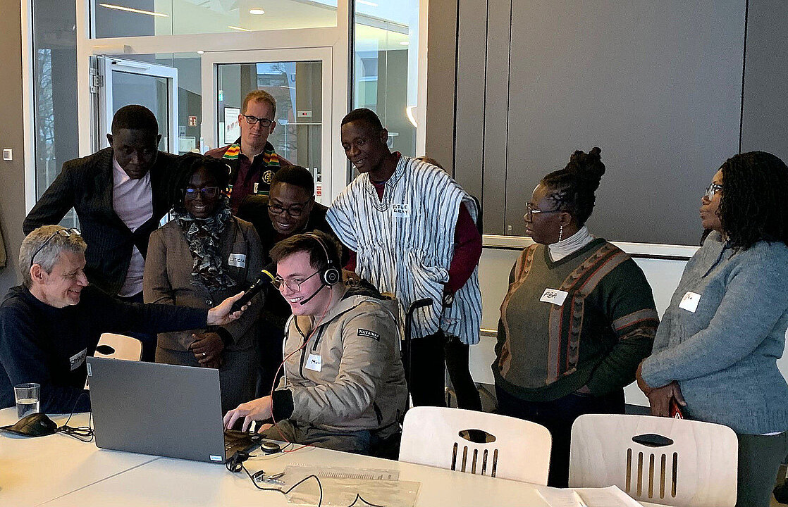 Mehrere Personen sitzen und stehen um einen Monitor und ein Mikrofon