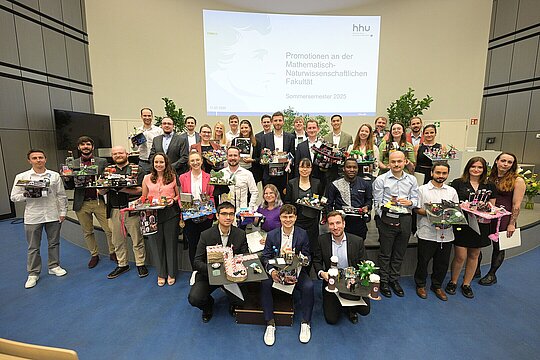 Gruppenbild aller Promovendinnen und Promovenden auf der Bühne des Hörsaals. Die Promovendinnen und Promovenden tragen ihre kreativ geschmückten Hüte auf den Armen.