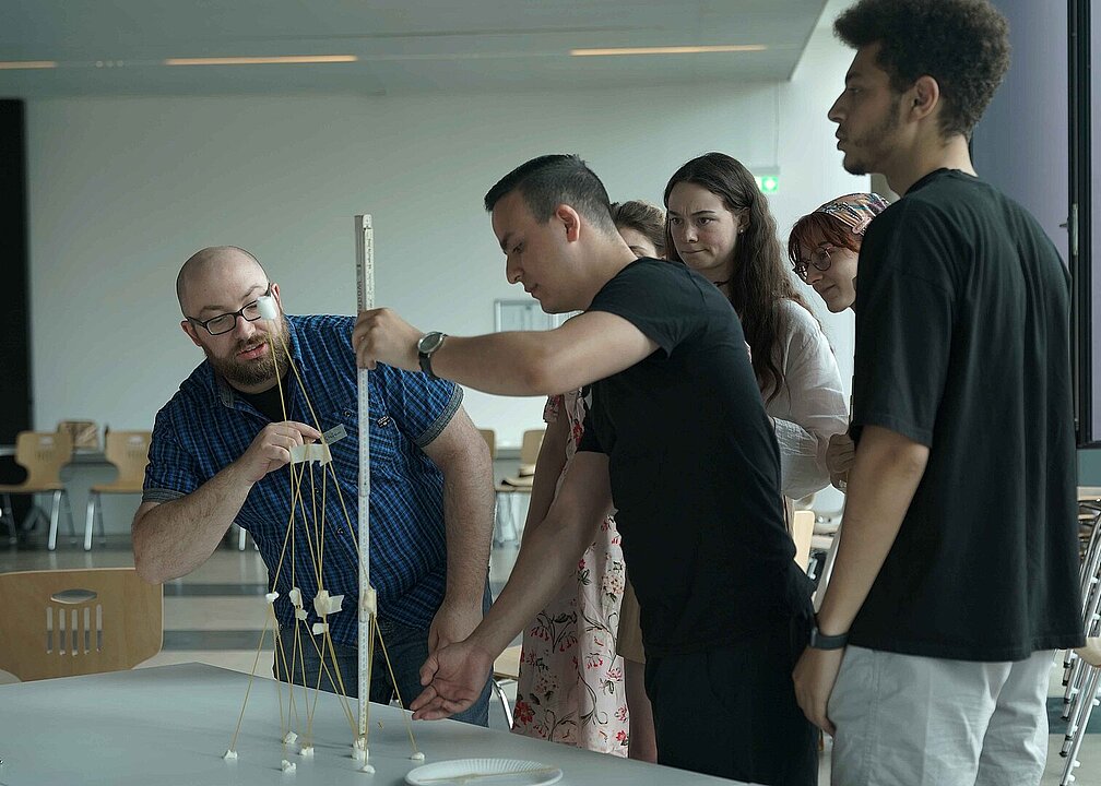 Mehrere personen stehen um einen Tisch und basteln mit einem Zollstock an einem Türmchen