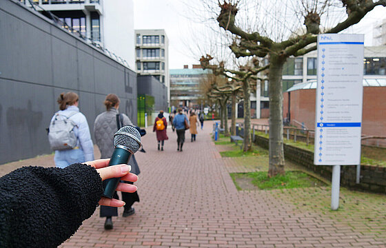 Eine ausgestreckte Hand hält ein Mikrofon. Im Hintergrund ist die leicht belebte Magistrale zu sehen.