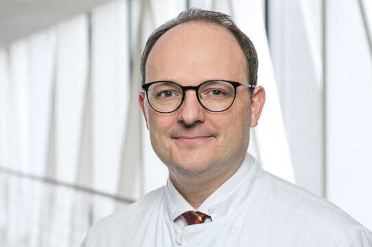 Portraitfoto von Prof. Dr. Sascha Dietrich. Er trägt eine runde, schwarze Brille, Halbglatze, einen weißen Kittel, ein weißes Hemd und ein rot-goldene Krawatte.
