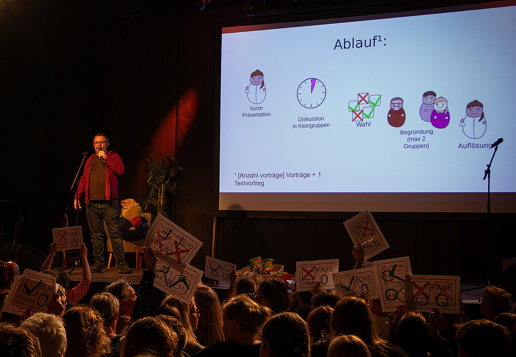 Moderator Tobias Löffler auf der Bühne, rechts von ihm eine Projektion mit der Erklärung des "Echt oder Fake"-Veranstaltungsablaufs.