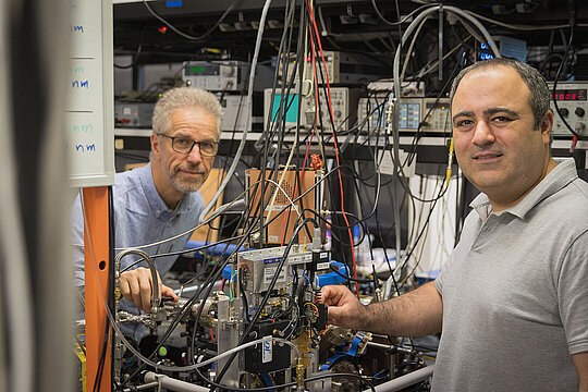 Die zwei Physiker stehen im Labor mit dem Messaufbau zwischen sich.