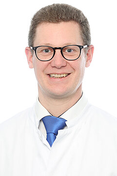 Portraitfoto von Prof. Dr. Neuerburg. Er trägt kurzes, dunkelgraues Haar, eine schwarze Brille, eine blaue Krawatte mit weißem Hemd und darüber einen weißen Kittel. Er lächelt.