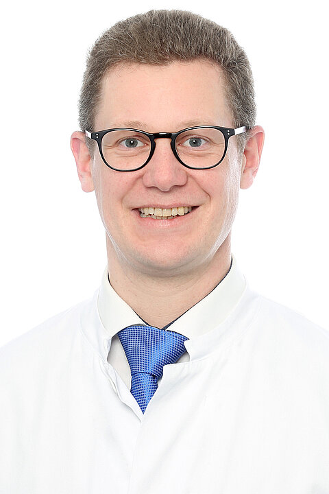Portraitfoto von Prof. Dr. Neuerburg. Er trägt kurzes, dunkelgraues Haar, eine schwarze Brille, eine blaue Krawatte mit weißem Hemd und darüber einen weißen Kittel. Er lächelt.