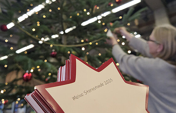 Sternchen vor einem Weihnachtsbaum im Hintergrund, an den gerade eine Person etwas hängt