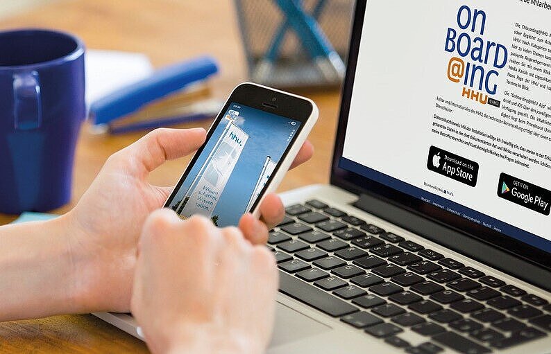 Eine Person hält ein Smartphone vor einem aufgeklappten Laptop in der Hand.