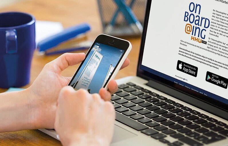 Eine Person hält ein Smartphone vor einem aufgeklappten Laptop in der Hand.