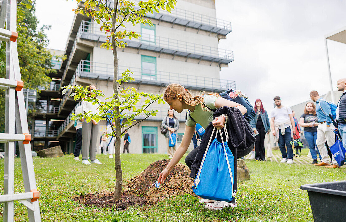 Eine junge Frau beugt sich zu einem frisch gepflanzten Baum hinunter, um einen Wunschzettel im Pflanzloch zu platzieren. Im Hintergrund sind weitere Azubis zu sehen.
