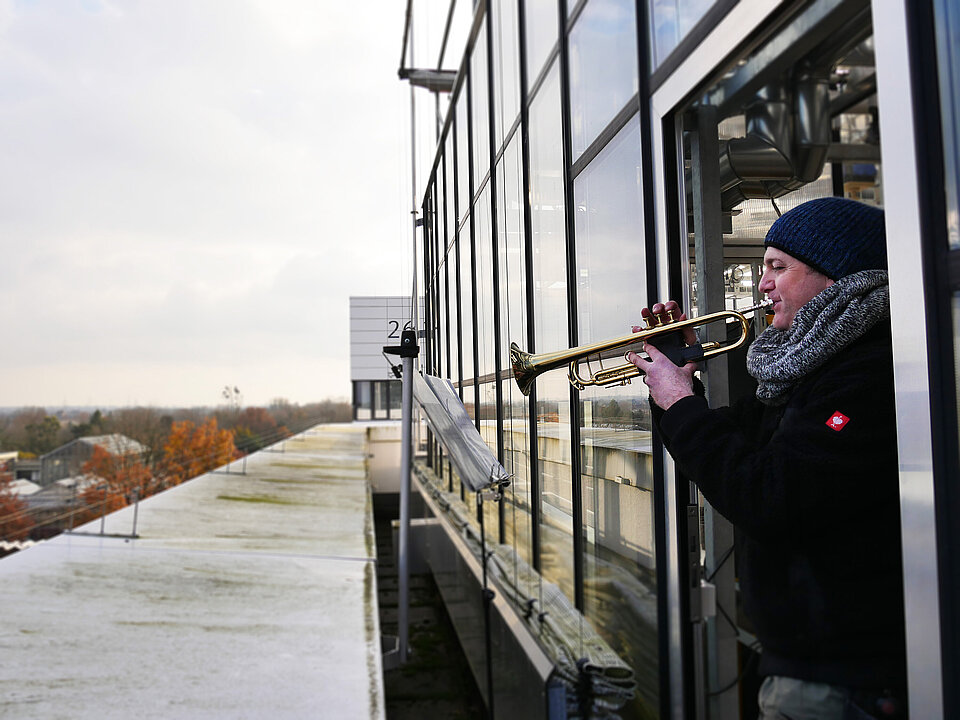In der offenen Gewächshaustür stehend bläßt Jochen Stappmanns in die Trompete, im Hintergrund der Botanische Garten der HHU und die Biologieneubauten.