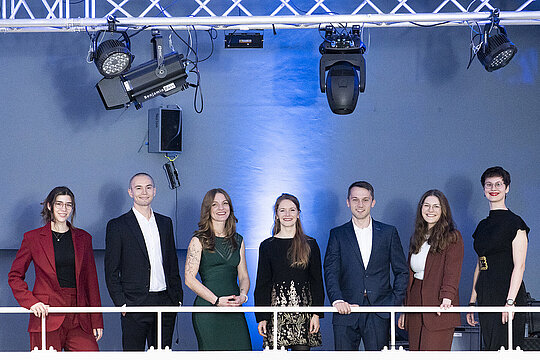 Gruppenbild mit acht nebeneinanderstehenden Personen vor blauer Wand hinter weißem Geländer.