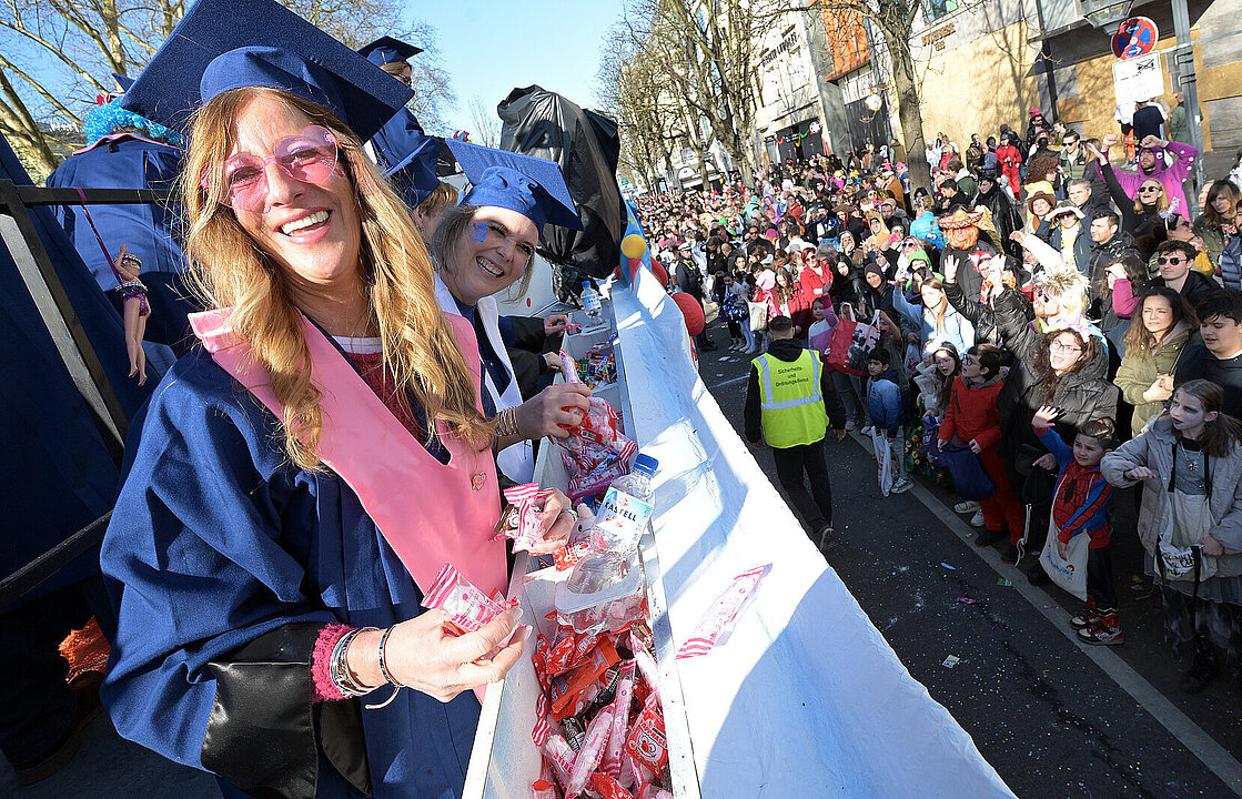 Die Wagenbesatzung lacht in die Kamera und wirft Kamelle in die Menge