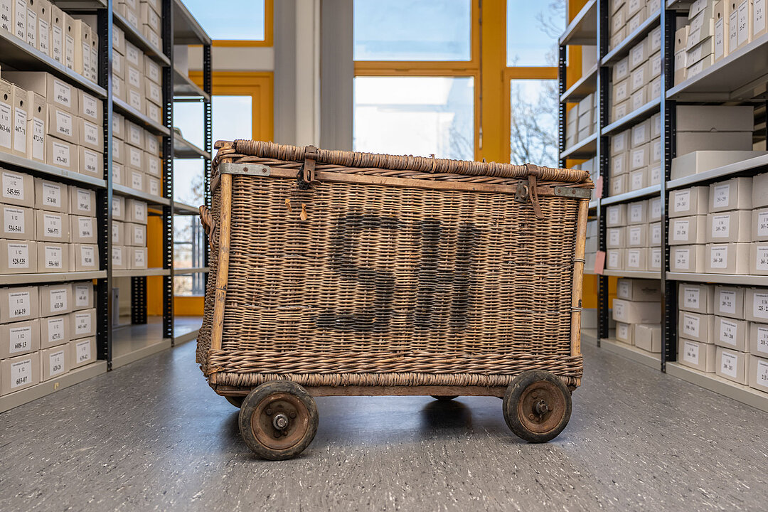Ein eckiger Wäschewagen aus geflochtenem Material steht zwischen Archivregalen