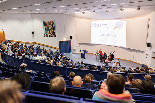 Aufnahme im Hörsaal mit Blick auf die Bühne, auf der ein Musiktrio spielt.