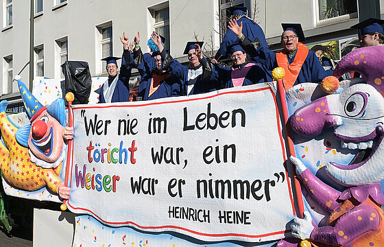 Karnevalswagen der HHU mit Besatzung. Das Heine-Zitat "Wer nie im Leben töricht war, ein Weiser war er nimmer" ist zu sehen.