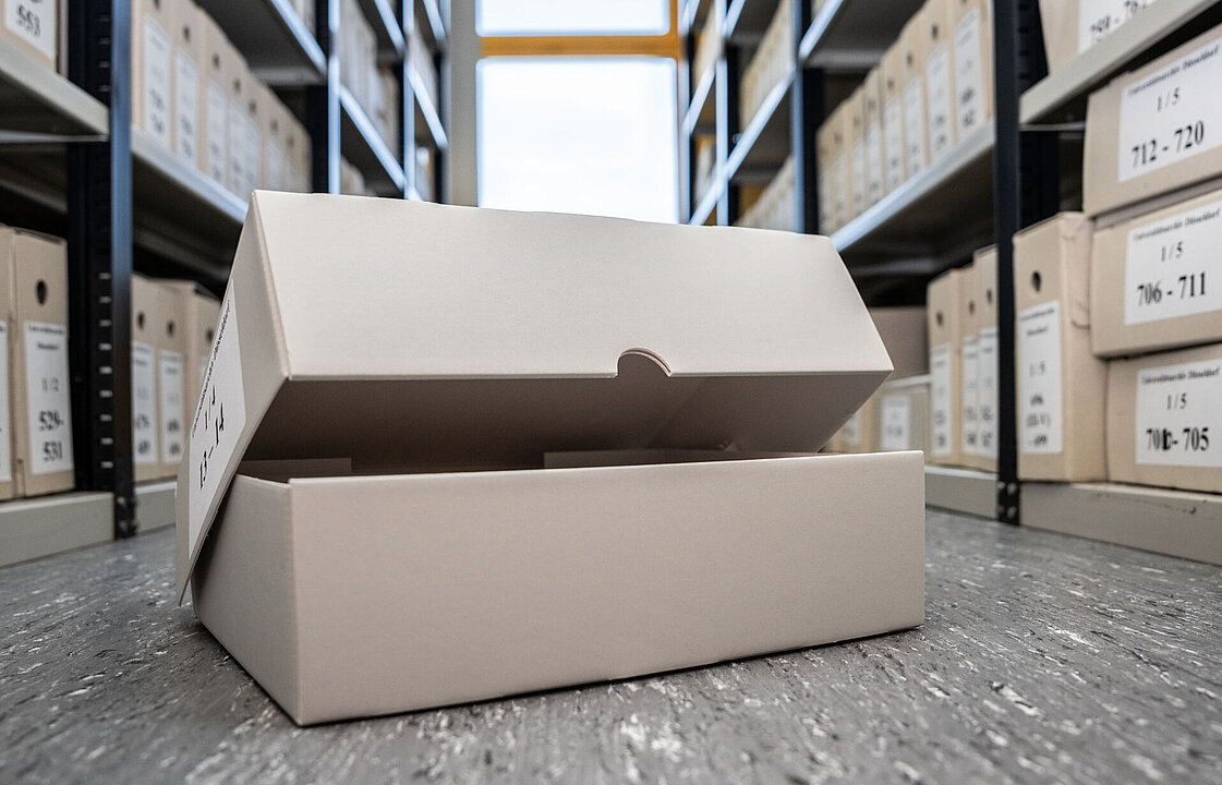 Ein grauer Karton steht halb geöffnet am Boden zwischen Archivregalen