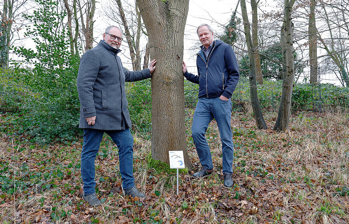 Zwei Personen neben dem Stamm einer Esche, dazwischen das Schild, das zur Baumpatenschaft verliehen wurde.