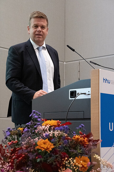 Matthias Heidmeier am Rednerpult stehend. Er hält grade eine Rede. Neben ihm ist der Blumenschmuck sichtbar.