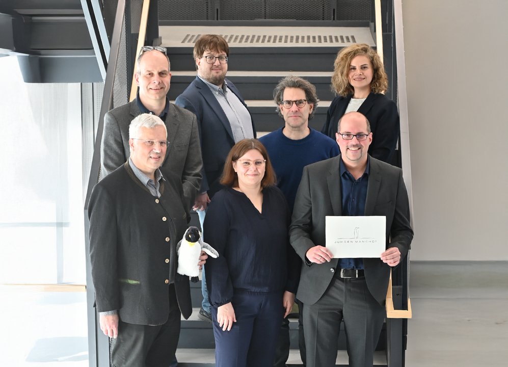 Gruppenbild mit zwei Damen und fünf Herren auf einer Treppe. Die Person links unten hält einen Stofftier-Pinguin in Händen, die Person rechts unten ein Schild mit dem aufgedruckten Logo der Jürgen Manchot Stiftung.