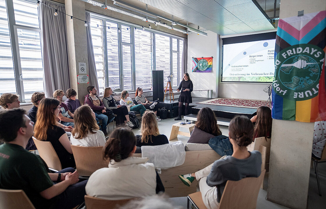 mehrere sitzende Personen hören einer vortragenden Person zu