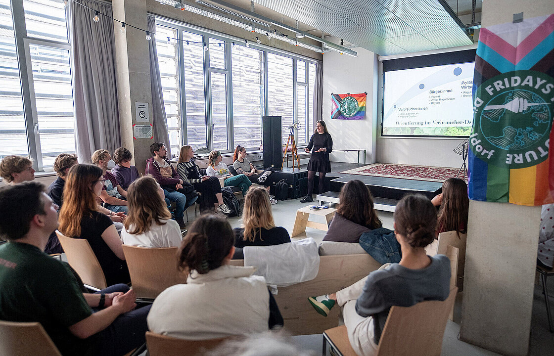 mehrere sitzende Personen hören einer vortragenden Person zu