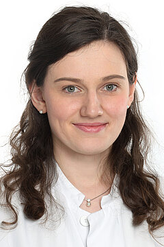 Portraitfoto von Prof. Dr. Laura Hinze. Sie trägt einen weißen Kittel, hat braune, lockige Haare in einem Seitenscheitel und trägt Perlenohrringe.