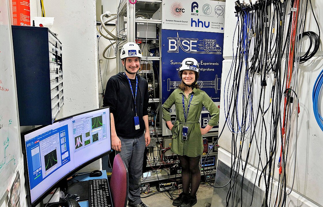 Zwei Personen mit Helm im Labor, links vorne ein Monitor, rechts viele Kabel, im Hintergrund eine Gerätewand mit HHU- und BASE-Logo.