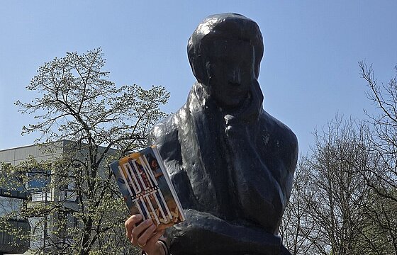 Die Heine-Statue am HHU-Campus und eine Hand, die daneben das Buch "Identitti" hochhält
