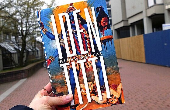 Eine Hand hält den Buchtitel "Identitti" hoch, im Hintergrund ist die Magistrale der HHU zu sehen.