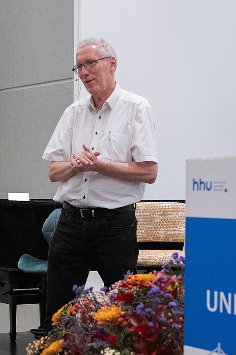 Prof. Dr. Stefan Wilm auf der Bühne im Haus der Universität stehend. Er trägt ein weißes Hemd und eine schwarze Hose. Neben ihm ist ein Blumenschmuck sichtbar. Seine Gestik und das Mikrofon zeigen, dass er grade in der Moderation ist.