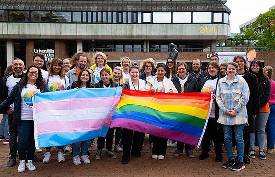 Eine Gruppe von Menschen steht mit einer Regenbogenflagge und einer bunten Flagge vor der Zentralbibliothek auf dem Campus der HHU: