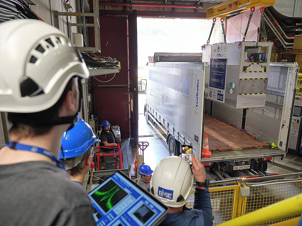 Ein Lkw steht in einer offenen Hallentür, mit Kran wird Apparatur auf die Ladefläche gehoben. Rechts Person mit Tablet.