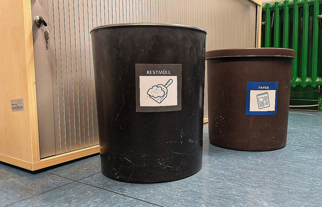 Zwei Sammelbehälter vor einem Schrank, darauf die Aufkleber "Restmüll" und "Papier"
