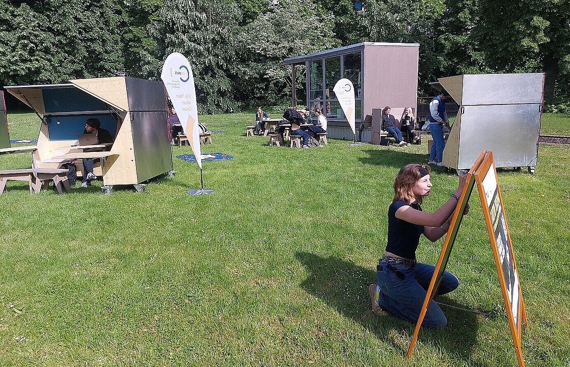 Eine Person schreibt etwas auf eine Tafel auf einer Wiese - dahinter Sitzmöglichkeiten aus Holz