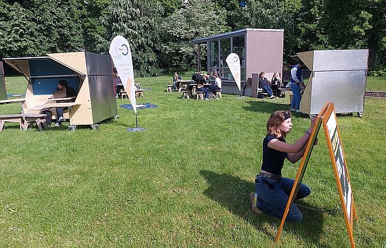 Eine Person schreibt etwas auf eine Tafel auf einer Wiese - dahinter Sitzmöglichkeiten aus Holz