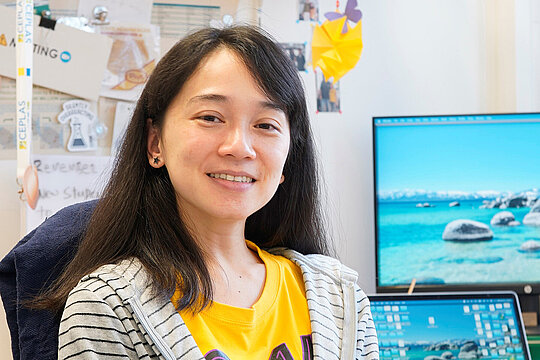 Portraitfoto von Dr. Eliza Loo in einem Büro, rechts sind zwei Monitore zu sehen.
