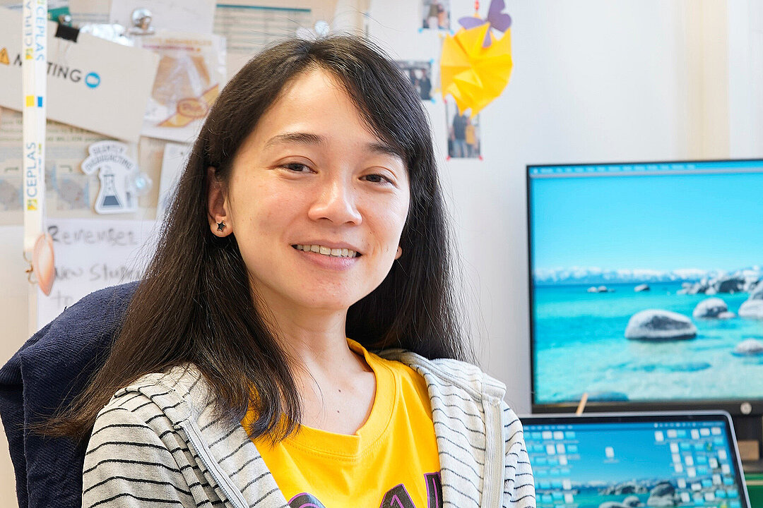 Portraitfoto von Dr. Eliza Loo in einem Büro, rechts sind zwei Monitore zu sehen.