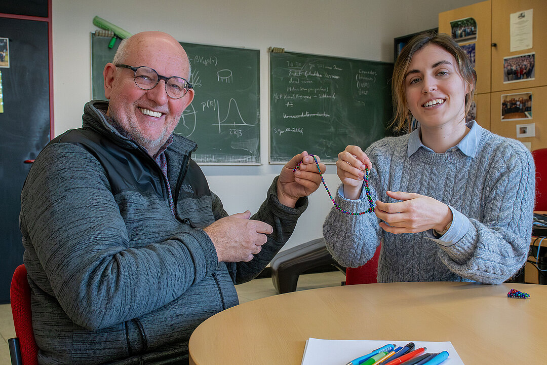 Prof. Dr. Hartmut Löwen (links) und Dr. Margaret Rosenberg in einem Büro. Sie halten gemeinsam eine Kette mit magnetischen Perlen in der Hand, die sich teilweise anziehen.