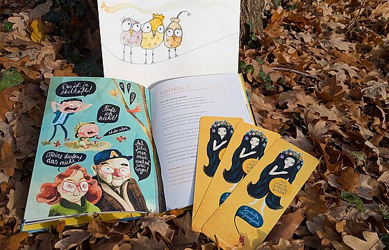 Ein aufgeschlagenes Buch und drei Lesezeichen lehnen an einem Baum.