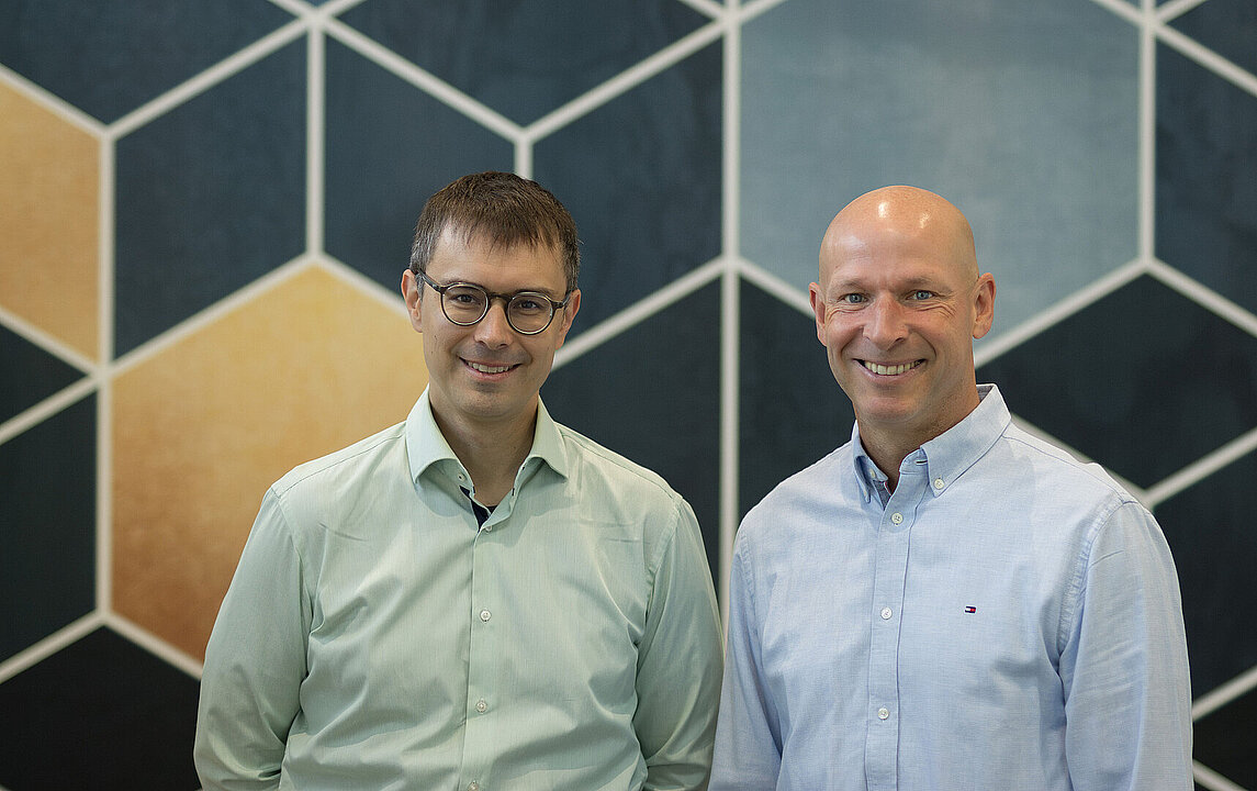 Die beiden Forscher von der HHU vor Wand mit geometrischen Mustern.