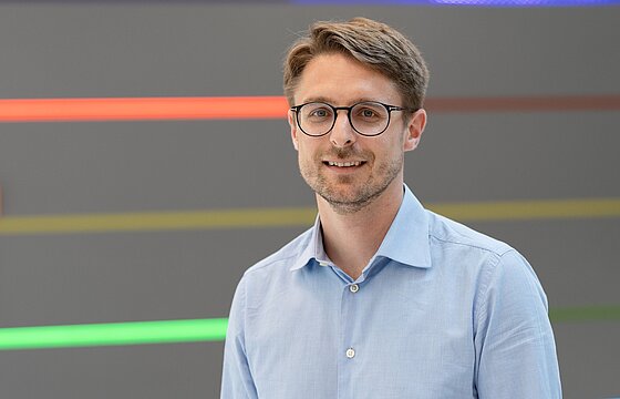 Portraitbild von Prof. Dr. Johannes Rebelein vor grauer Wand, die farbige horizontale Streifen zeigt.