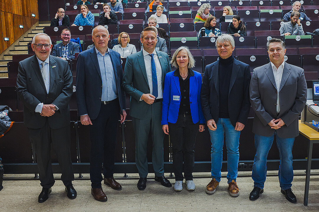 Gruppenbild von sechs Personen, im Hintergrund ein besetzter Hörsaal.