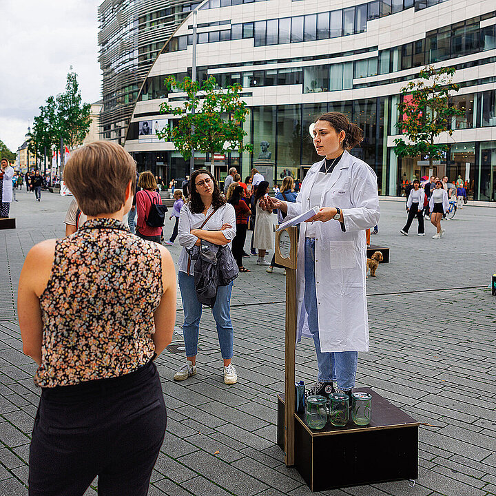 Eine Sprecherin im weißen Kittel steht bei der Veranstaltung 2023 auf einer Holzkiste auf dem Schadowplatz in Düsseldorf und berichtet Zuhörerinnen von ihrer Forschung.