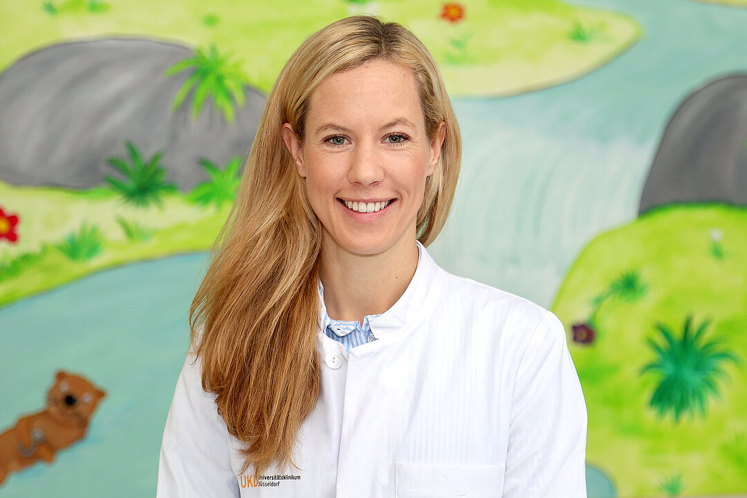 Portraitfoto von Dr. Röper, sie trägt einen weißen Kittel und steht vor einer bunt bemalten Wand in der Kinderklinik des UKD.