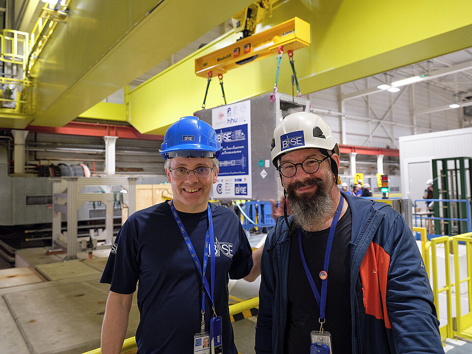 Zwei lächelnde Männer mit Helmen vor einem großen Traversenkran.