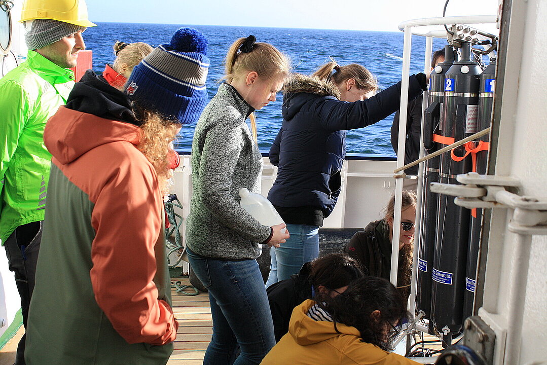 Auf dem Schiffsdeck arbeitet eine Gruppe von Wissenschaftlern und Studierenden an dem Wasserprobennehmer, der ins Wasser gelassen werden soll.