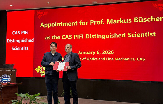 Zwei Personen vor Wand mit Präsentation; Prof. Büscher hält eine Urkunde vor sich.