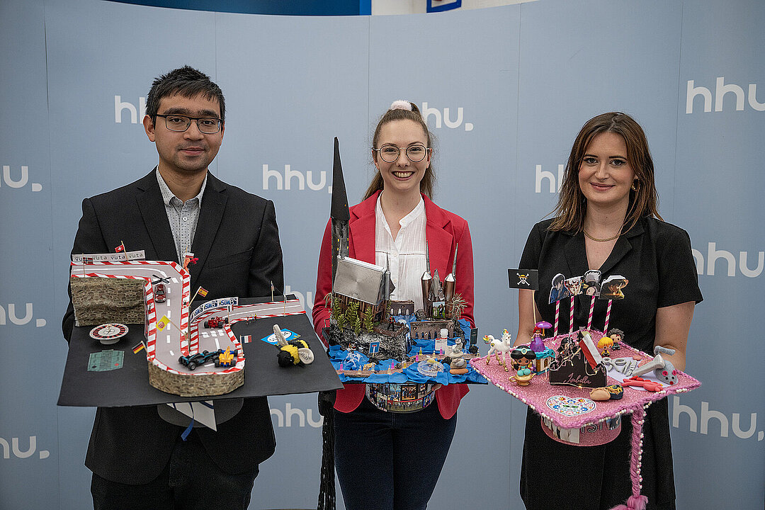 Die drei Preisträgerinnen und Preisträger auf einem Gruppenbild. Sie tragen ihre kreativ geschmückten Hüte auf dem Arm. Von links nach recht: Herr Fabian Vu mit einem Formel-1-Hut, Frau Patricia Kleimann mit einem Hogwarts-Hut, Frau Isabella Maria Solga-Kamp mit einem Hut ganz in rosa.